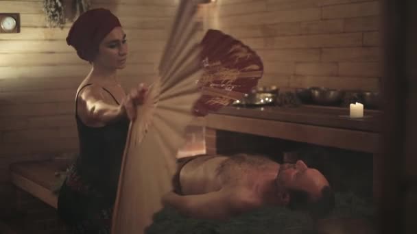 Женщина Проводит Ритуал Исцеления Китайским Веером Мужчины Человек Лежит Еловых — стоковое видео