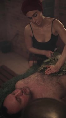 Bir kadın süpürgelerin yardımıyla bir erkek için banyo ritüeli gerçekleştiriyor. Bir adam ladin dallarına uzanır..