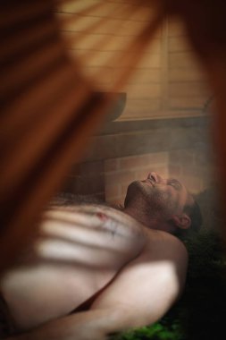 Bir saunadaki ladin dallarına uzanan bir adam, Çinli bir vantilatörün içinden ışık sızıyor. İyileşme ritüeli.