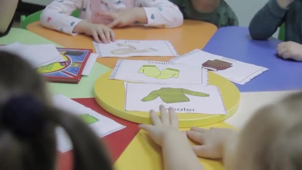 小さな子供たちのグループは幼稚園で冬服の英語名を学びます 先生は丸いテーブルの上に紙のシートを置いてイラストをよく見る — ストック動画