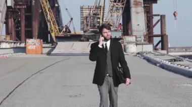 İnşaat alanında telefonla konuşan ve kameranın önünde yürüyen bir iş adamı.