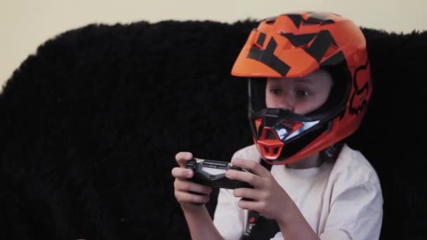 男孩戴着橙色头盔坐在沙发上玩游戏机 — 图库视频影像