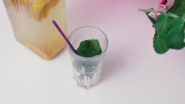 Kadın taze limonata yapmak için beyaz masaya yeşil nane yapraklı bardağa su döküyor. Ev hanımı kavanozu limonla yakın çekimde tutar.