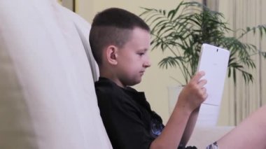 Çocuk tablette oyun oynuyor, beyaz tüylü bir tavşan kanepede zıplıyor. Yeşil çalı kapat