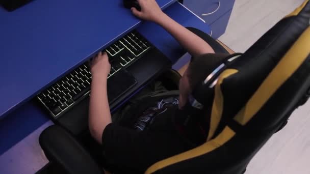 头戴耳机的男孩坐在靠近橙色头盔的办公桌前玩电子游戏 上瘾的青少年喜欢在家中近距离观看电脑上的赛车 — 图库视频影像