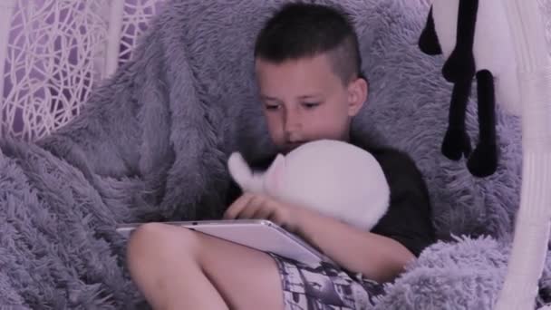 Boy Senta Uma Cadeira Ovo Joga Tablet Segura Coelho Branco — Vídeo de Stock
