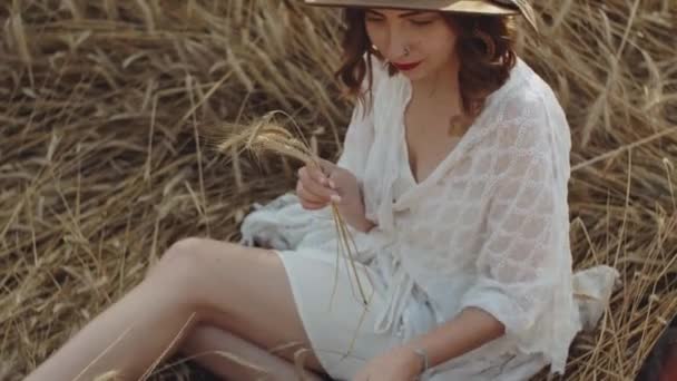 Uma mulher bonita em um vestido branco e chapéu marrom que senta-se em um campo entre spikelets e joga com spikelets, campos da Ucrânia, paisagem bonita — Vídeo de Stock