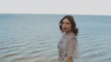 Kırmızı dudaklı, kıvırcık saçlı, kameraya gülümseyen çekici bir kadın. Mavi deniz, dalgalar ve mavi gökyüzü..