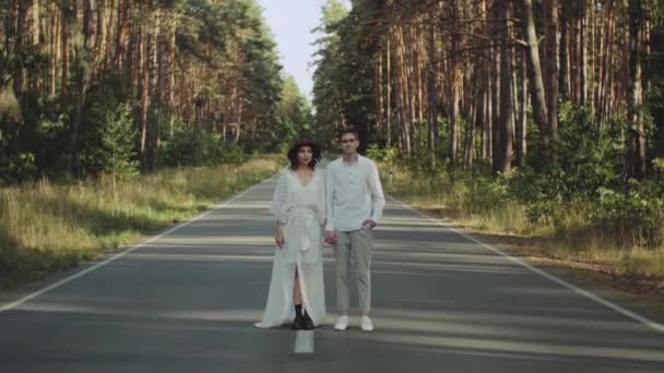 Egy gyönyörű hipszter pár áll az út közepén kézen fogva és csókolózva. Út fut keresztül az erdőn, napos idő. — Stock videók