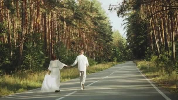 Влюбленные хипстеры прогуливаются в красивом месте, держатся за руки, мужчина и женщина обнимаются и наслаждаются общением на пустой дороге через живописный сосновый лес — стоковое видео