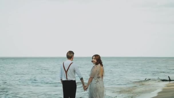 Hermosa pareja joven enamorada caminando por la playa mientras las olas inundan la playa disfrutando del paseo. Pareja enamorada cogida de la mano. — Vídeo de stock