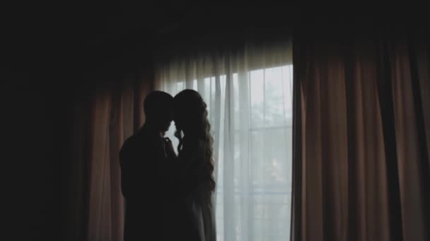 Egy szerelmes férfi és nő sziluettje ölelkezik az ablaknál. Közepes felvételű sziluett friss házaspárról. Házasság, romantikus légkör. — Stock videók