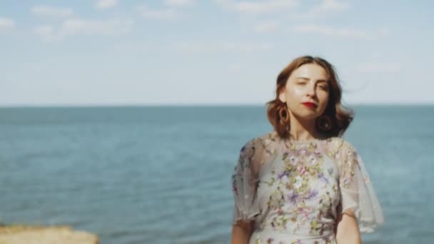 Attraktive Frau mit roten Lippen berührt ihre lockigen Haare zu Fuß, dreht sich um und blickt in die Kamera, Zeitlupe. blaues Meer mit Wellen und blauem Himmel im Hintergrund. — Stockvideo