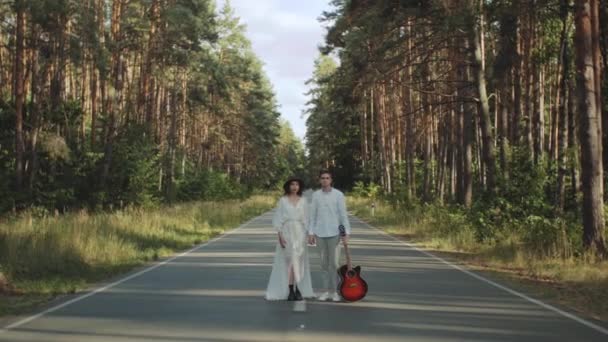 A beautiful hipster couple stands in the middle of the road holding hands and kissing slow motion. Road running through the forest, sunny weather. — Stok video