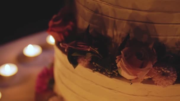 Grote mooie bruidstaart versierd met bloemen in de avond in de buurt van de boog close-up, slow motion. kaarsen op achtergrond. — Stockvideo