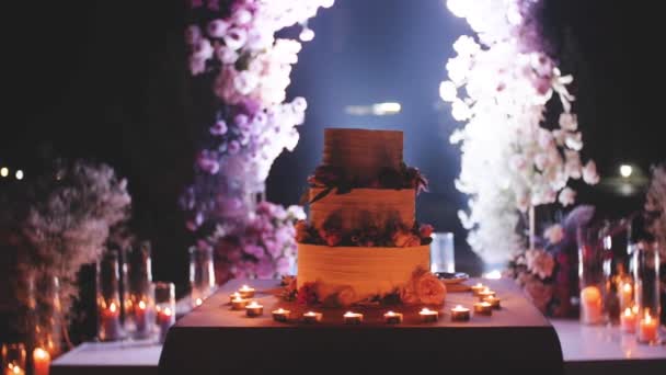 Große schöne Hochzeitstorte, abends in der Nähe des Bogens mit Blumen dekoriert, mittlerer Schuss, Zeitlupe. Kerzen rund um den Kuchen. — Stockvideo