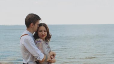 Mutlu çift kucaklaşması, orta boy. Adam kıza arkadan sarılıyor ve kadın kameraya bakıyor, yavaş çekim, arka planda deniz var, rüzgar kadın saçlarını büyütüyor. 4k