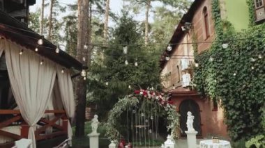 Kırmızı çiçekli düğün kemeri ve beyaz sandalyeler, restoranın yakınındaki törenin üzerine lamba çelengi. Boho-şık düğün tarzı. Yüksek kaliteli FullHD görüntüler