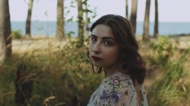 Kırmızı dudaklı çekici bir kadın kıvırcık saçlarına dokunur, yürür, döner ve kameraya bakar, alçak hareket. Mavi deniz, dalgalar ve mavi gökyüzü. Yüksek kalite 4k görüntü