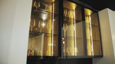 Black cupboard with glass wall in the kitchen with crockery and clear glasses with lighting. Clean wine glasses in cabinet. Contemporary Design Style. Kitchen Room. Slow motion. 4k footage