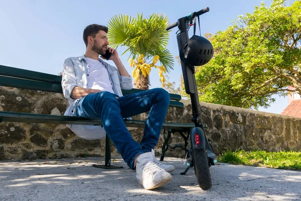Genç adam akıllı telefonda elektrikli scooterının yanında konuşuyor.