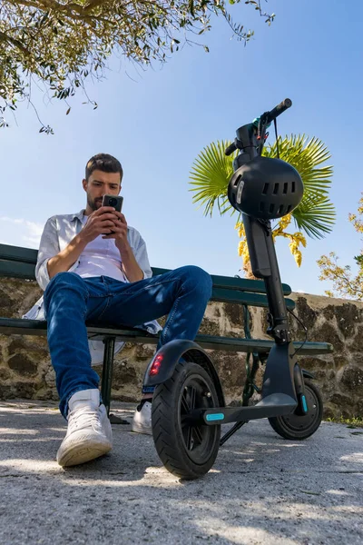 Genç adam akıllı telefonuna bakar. Parktaki bankta elektrikli scooterının yanında oturur.