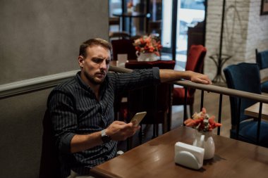 Adamın biri kafedeki bir masada bir koltuğa oturup telefona bakıyor.