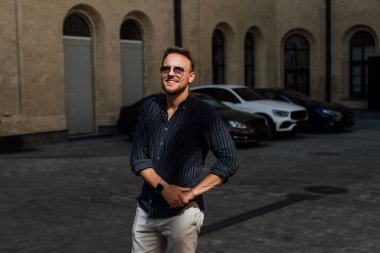 Gömlekli, gözlüklü ve pantolonlu bir adam park halindeki arabaların önünde duruyor.