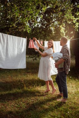 Beyaz elbiseli hamile bir kız kocasıyla bahçede bebek kıyafetleri asıyor.