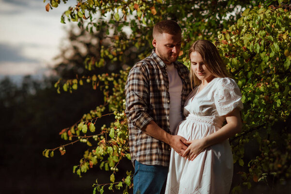 A pregnant girl in a white dress and her husband in a shirt near a tree in the garden
