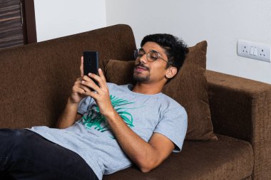 Genç bir çocuk kanepede uzanmış akıllı telefon kullanıyor ve gülümsüyor.