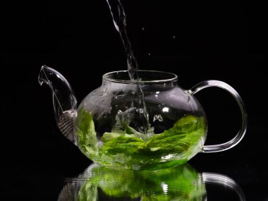 Siyah zemin üzerinde cam bir çaydanlığa sıcak su dökmek, yeşil nane çayı. Bitki çayı ve sağlıklı içecek konsepti.