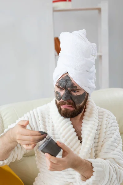 Barbudo hombre divertido que se divierte con una máscara cosmética en su cara hecha de arcilla negra. Cuidado de la piel de los hombres, humor y spa en el hogar concepto — Foto de Stock