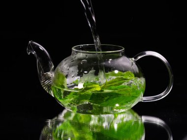 Siyah zemin üzerinde cam bir çaydanlığa sıcak su dökmek, yeşil nane çayı. Bitki çayı ve sağlıklı içecek konsepti.