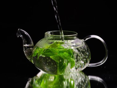 Siyah zemin üzerinde cam bir çaydanlığa sıcak su dökmek, yeşil nane çayı. Bitki çayı ve sağlıklı içecek konsepti