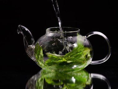 Siyah zemin üzerinde cam bir çaydanlığa sıcak su dökmek, yeşil nane çayı. Bitki çayı ve sağlıklı içecek konsepti.