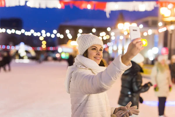 Mujer joven feliz en invierno en la pista de hielo tomando fotos en el teléfono inteligente, selfie. —  Fotos de Stock