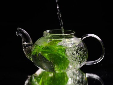 Siyah zemin üzerinde cam bir çaydanlığa sıcak su dökmek, yeşil nane çayı. Bitki çayı ve sağlıklı içecek konsepti.