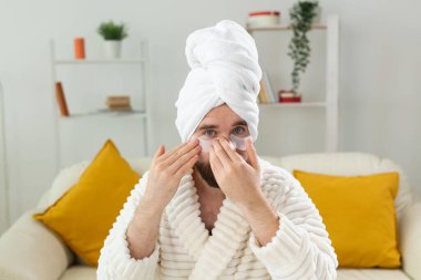 Evde gözlerinin altında yamalar olan genç bir adam. Erkekler için kozmetoloji kavramı. Evde spa, vücut ve cilt bakımı