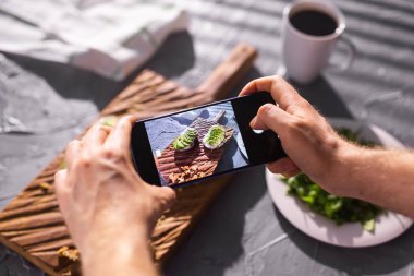 Eller, masada duran iki güzel sağlıklı ekşi krema ve avokado sandviçinin resimlerini akıllı telefondan çeker. Sosyal medya ve gıda kavramı