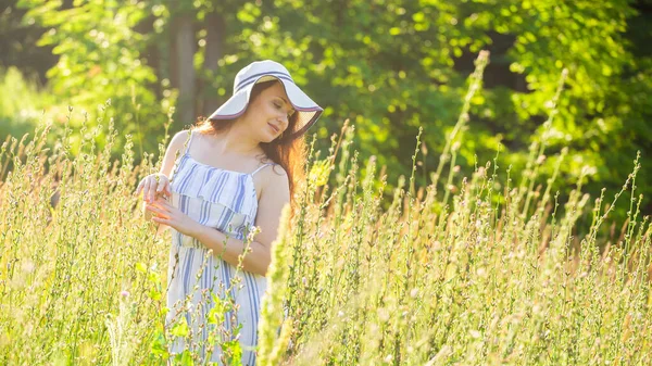 Joyeux jeune femme aux cheveux longs en chapeau et robe tire ses mains vers les plantes tout en marchant dans la forêt d'été par une journée ensoleillée. Bannière avec espace de copie — Photo