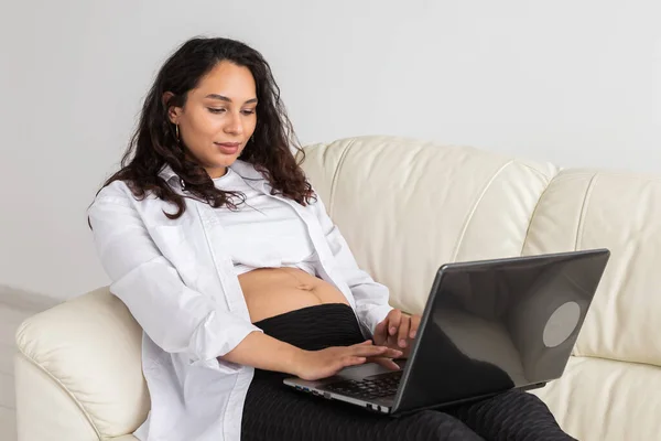 Gravid kvinna som använder laptop när hon sitter på en soffa i vardagsrummet hemma — Stockfoto