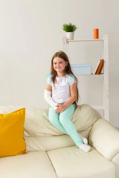 Enfant avec un plâtre sur un poignet ou un bras cassé souriant et s'amusant sur un canapé. Attitude positive, récupération et concept enfant. — Photo