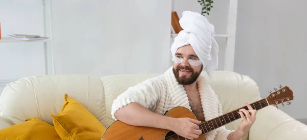 Un jeune homme avec des patchs sous les yeux joue à la guitare à la maison. Concept de cosmétologie pour homme et musicien. Spa à la maison, musique et soins de la peau — Photo