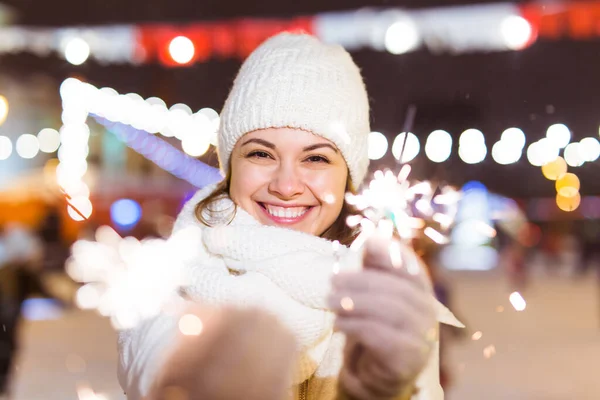 Giovane bella donna in maglia cappello e sciarpa in piedi in città con luce bengala, scintillante. Concetto celebrazione e Natale. — Foto Stock