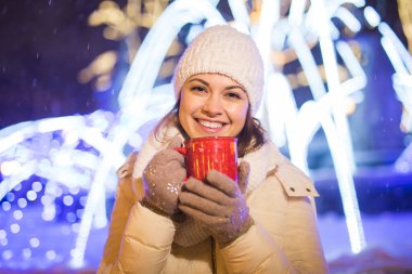 Noel tatili. Kışın sıcak bir fincan kahveyle sıcak kıyafetler içinde gülümseyen güzel bir kadın. Şehir gecesi.