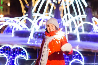 Kış örgü giysileri giymiş, dışarıda kar altında maytap tutan gülümseyen genç bir kadın. Noel tatili.