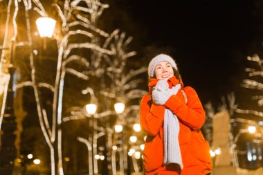 Arka planda kışlık kıyafetleri olan neşeli, eğlenceli genç bir kadın. Şehir ışıkları aydınlanıyor. Noel ve kış tatili konsepti.