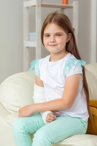 Barn med gips på bruten handled eller arm som ler och har roligt på soffan. Positiv attityd, återhämtning och barnkoncept. — Stockfoto