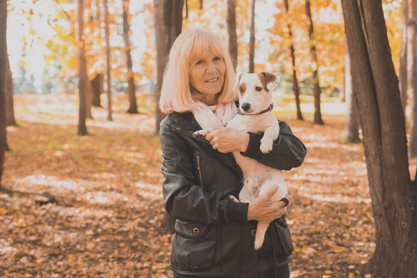 Senior lachende vrouw die haar hond knuffelt in het herfstpark. Actief ouder worden en huisdier concept. — Stockfoto